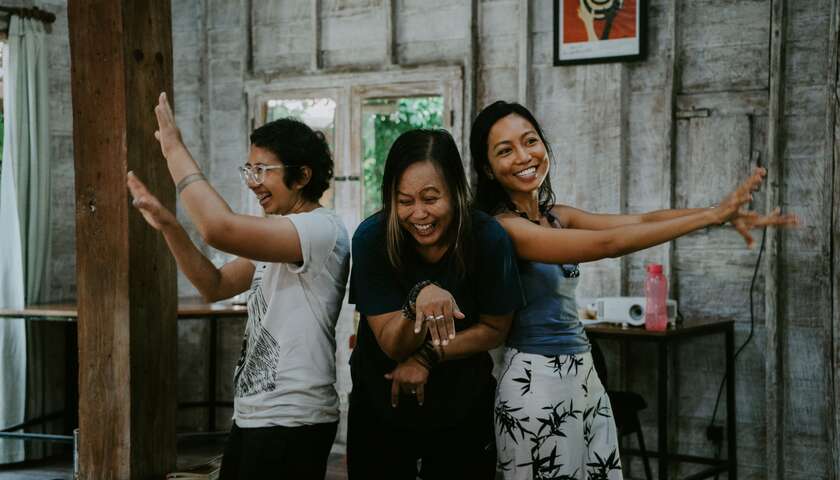 Three workshop participants stand in a circle with their backs to each other, aise their arms in the air and laugh.
